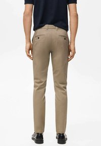 Pantaloni chino beige con taglio slim, dotati di due tasche posteriori con chiusura a bottone, indossati con scarpe nere. Il tessuto sembra liscio e resistente.