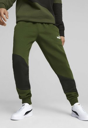 Person trägt grüne und schwarze Puma-Sweatpants und weiße Puma-Sneakers, steht mit gespreizten Beinen vor einem einfarbigen Hintergrund.