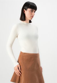 Jeune femme avec des cheveux foncés et raides portant un haut à manches longues blanc à côtes et une jupe en velours côtelé marron, se tenant devant un fond blanc uni.