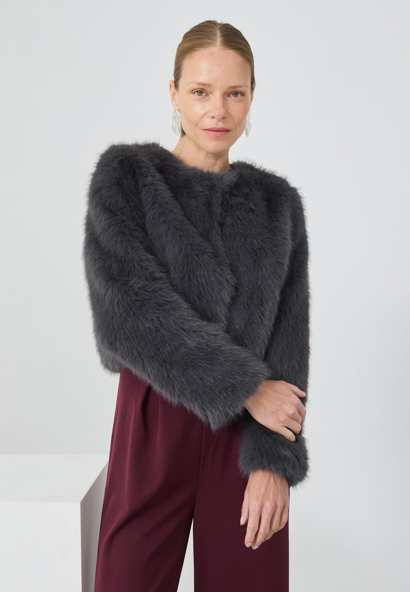 Mujer con el cabello recogido hacia atrás que lleva pendientes de plata, una chaqueta de piel sintética gris oscuro y pantalones de talle alto burdeos frente a un fondo simple.