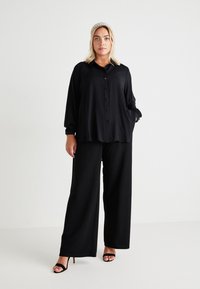 Black long-sleeve button-up shirt with a loose fit, featuring a collar and gathered cuffs, paired with wide-leg black trousers and strappy heels.