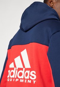 Sweatshirt com capuz apresentando uma parte superior azul-marinho e uma parte inferior vermelha, com um destacado logo branco da Adidas na frente. Textura de tecido suave.