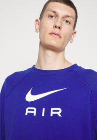Ljubičasti dukserica s bijelim Nike swooshom i natpisom "AIR", s okruglim izrezom i rebrastim manžetama. Vidljiva meka tekstura tkanine.