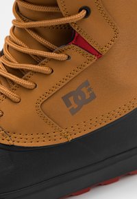 Bruine leren laars met zwarte rubberen zool, voorzien van oranje veters, gestikte accenten en een zichtbaar logo aan de zijkant.