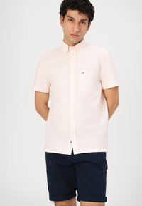Camisa de botones de manga corta en rosa claro con un tejido texturizado, que presenta un bolsillo en el pecho y un pequeño logo como detalle. Llevada con pantalones cortos de color navy.