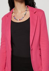 Rosa vävd kavaj med skräddarsydd siluett, matchad med en svart linne. Utrustad med ett färgglatt pärlhalsband med olika former och färger.