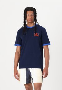 Marinblå bomulls t-shirt med korta ärmar, med en kontrasterande blå halsringning och ärmkanter, samt en orange broderad logotyp-patch.