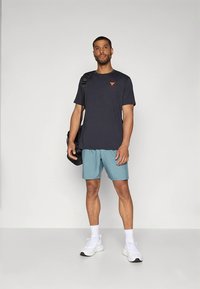 Donkergrijs heren T-shirt met oranje logo, gecombineerd met lichtblauwe korte broek. Witte sneakers, zwarte schoudertas, neutrale achtergrond.