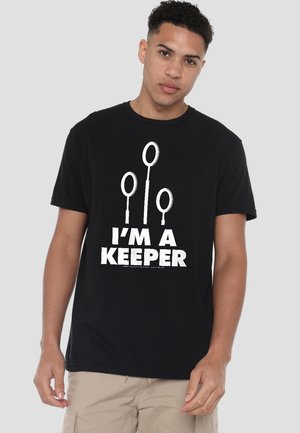 Young man wearing a black T-shirt with three Quidditch hoops and the text "I'M A KEEPER" in white, standing against a plain background.