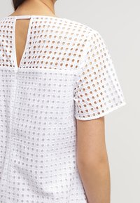 White short-sleeve top featuring a textured grid pattern, open back with a small slit, and a relaxed fit design.