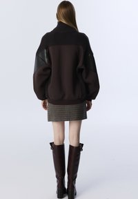 Vrouw in een oversized donkerbruin jack met leren patches, een geruite mini-jurk en bruin leren kniehoge laarzen, staand tegen een witte achtergrond.