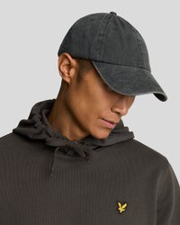 Casquette de baseball grise avec une texture douce, visière courbée et un bouton sur le dessus, portée avec un sweat à capuche sombre arborant un petit logo jaune.