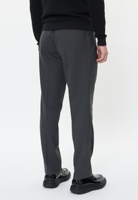 Šedé kalhoty na míru s hladkým texturem, s rovnými nohavicemi a diskrétními sklady. Nosené s černými botami, zobrazený zadní pohled.