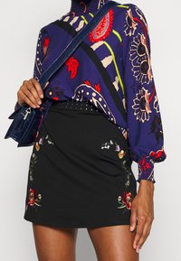 Paars gebloemd blouse met rode en groene accenten, gecombineerd met een zwarte rok met kleurrijke bloemborduursels en een gestudeerde tailleband.