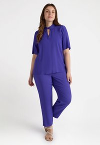 Lila Zweiteiler, bestehend aus einer kurzärmeligen Bluse mit knöpfbarem Kragen und einem Schlüssellochdetail sowie passenden, weit geschnittenen Hosen.