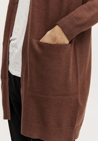 Cardigan tricotat maro cu un croi lejer, mâneci lungi, manșete ribbate și un buzunar frontal. Textura este moale, cu un luciu subtil.