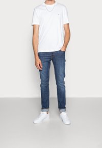 Giovane uomo che indossa una maglietta bianca e jeans blu, in piedi contro uno sfondo neutro, con le mani nelle tasche e sneakers bianche.