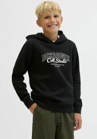 Smaidošs blondīns puika, ģērbies melnā Jack & Jones kapucē un zaļās biksēs, stāv ar vienu roku kabatā pret vienkāršu fona.