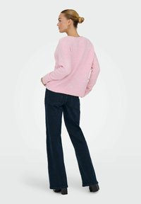 Roze gebreide trui met geribde zoom, gecombineerd met donkerblauwe, uitlopende jeans. De stof oogt zacht en gestructureerd, wat de casual stijl versterkt.