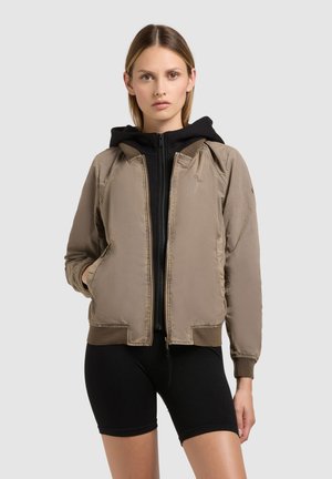 Ung kvinde iført en beige bomberjakke over en sort hættejakke med lynlås og sorte cykelshorts, stående foran en ensfarvet baggrund.