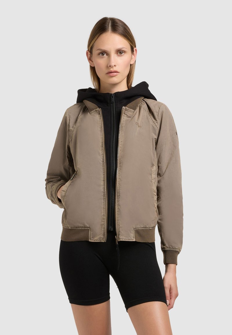 Jeune femme portant une veste bomber beige par-dessus un sweat à capuche noir zippé et un short de cyclisme noir, debout devant un fond uni.