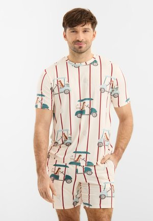 Mies, joka pukeutunut samansävyiseen kermaan olevaan paitaan ja shortseihin, joissa on punaiset raidat ja sininen golfkärrykuvio, seisoo yhdellä kädellä taskussa.