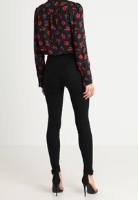 Blusa negra con flores rojas y verdes, jeans ajustados negros y zapatos de tacón negros. La blusa tiene un escote fruncido y mangas largas.