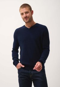 Marineblauer, gestrickter Pullover mit V-Ausschnitt und gerippter Textur, mit langen Ärmeln und taillierter Passform. Getragen mit dunklen Jeans.