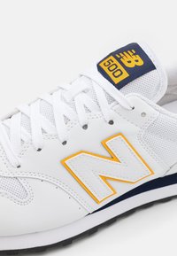 Baskets blanches en mesh et cuir, avec un grand logo "N" jaune, des accents bleu marine et des lacets blancs.