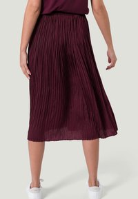 Gonna midi plissettata in un profondo bordeaux, tessuto leggero con pieghe verticali. Vita elasticizzata e silhouette a trapezio, abbinata a sneaker bianche.