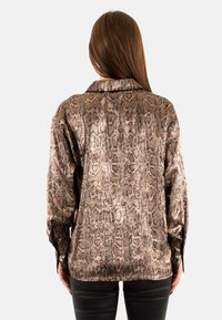 Blusa marrón con estampado de serpiente, de textura suave, que presenta mangas largas y un cuello. El diseño incluye un brillo sutil y un corte relajado.