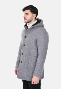 Cappotto grigio in lana con cappuccio, caratterizzato da bottoni neri, tasche laterali e una texture morbida. Indossato sopra un maglione chiaro in un ambiente neutro.