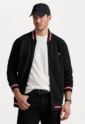 Giacca nera con zip e polsini a costine rossi e bianchi; indossata sopra una maglietta bianca, abbinata a jeans neri e un cappellino coordinato.