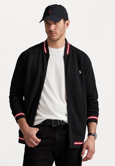 Giacca nera con zip e polsini a costine rossi e bianchi; indossata sopra una maglietta bianca, abbinata a jeans neri e un cappellino coordinato.