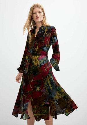 Femme blonde portant une robe à manches longues, col et ceinture, avec un motif abstrait coloré en rouge, jaune, bleu et vert.