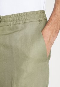 Homme portant un pantalon décontracté vert clair avec taille élastique, main reposant dans la poche avant droite, bras et torse visibles en chemise blanche.