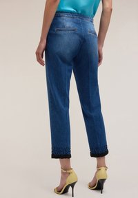 Woman wearing blue cropped jeans with black bead embellishments on cuffs and yellow high-heeled sandals, standing with back facing camera.