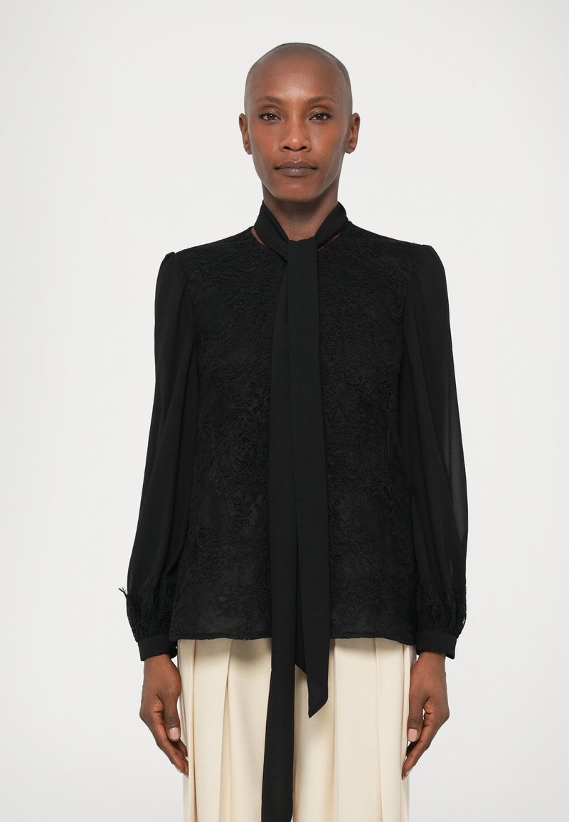 Melna blūze ar teksturētu mežģīņu priekšpusi, garām piedurknēm un auduma saitēm ap kaklu. Ievērojamas elastīgas aproces un brīva piegriezuma.