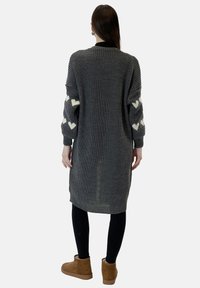 Cardigan largo gris de punto con tela texturizada, mangas anchas con patrones de corazones blancos y aberturas laterales. Lleva puesto sobre leggings negros y botas marrones.