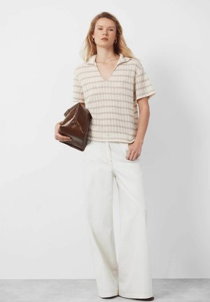 Frau trägt ein beige gestreiftes kurzärmeliges Oberteil und weite weiße Hosen, hält eine große braune Leder-Clutch.