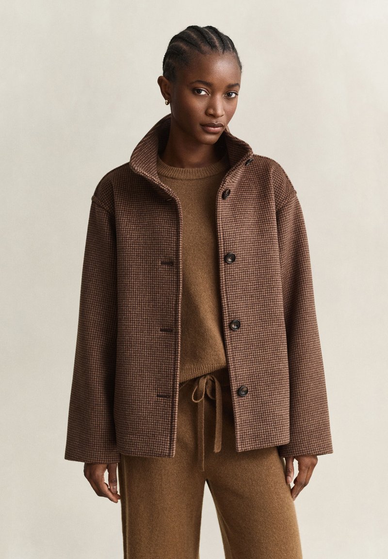 Femme portant une veste marron à carreaux boutonnée sur un pull marron assorti et un pantalon à cordon, debout devant un fond clair uni.