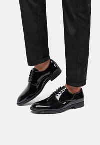 Scarpe eleganti in pelle verniciata nera con finitura liscia, punta arrotondata e design con sei occhielli per i lacci, abbinate a pantaloni neri alla caviglia.