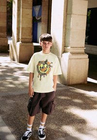 Light yellow T-shirt with a camo smiley graphic, paired with dark cargo shorts, white socks with yellow stripes, and black sneakers.