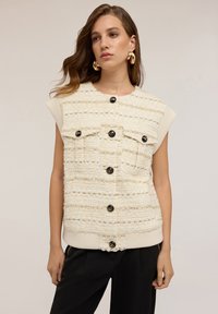 Una donna con capelli castani ondulati indossa un gilet senza maniche in tweed crema con bottoni neri e orecchini a cerchio dorati, abbinato a pantaloni neri.