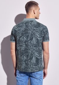 Camisa polo verde de manga corta con un patrón texturizado de hojas, costuras detalladas y un cuello clásico. Combinada con jeans azules.