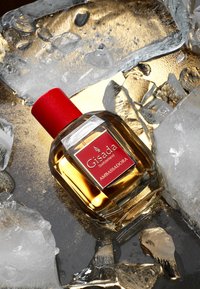 Butelka perfum Gisada Ambassadora z prostokątnym szklanym korpusem, złotym płynem, czerwoną nakrętką i czerwoną etykietą, umieszczona na lodzie na złotym tle.