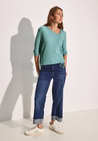 En turkos ribbad tröja med vid halsringning och trekvartsärmar, tillsammans med avslappnat passande mörk denimjeans och vita sneakers.