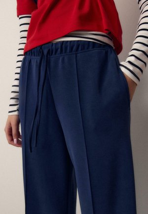 Person trägt marineblaue Jogginghosen mit Kordelzug, rotes Oberteil und ein schwarz-weiß gestreiftes Langarmshirt, eine Hand in der Tasche.