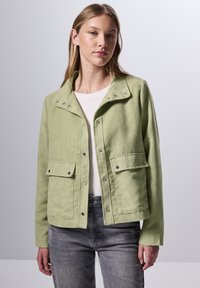 Chaqueta de pana verde claro con broches a presión, dos bolsillos frontales y un cuello. Llevada sobre una camisa blanca y unos jeans grises.