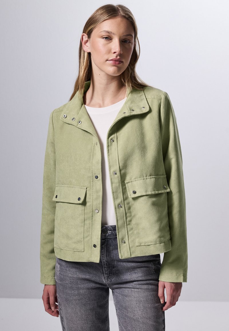 Chaqueta de pana verde claro con broches a presión, dos bolsillos frontales y un cuello. Llevada sobre una camisa blanca y unos jeans grises.